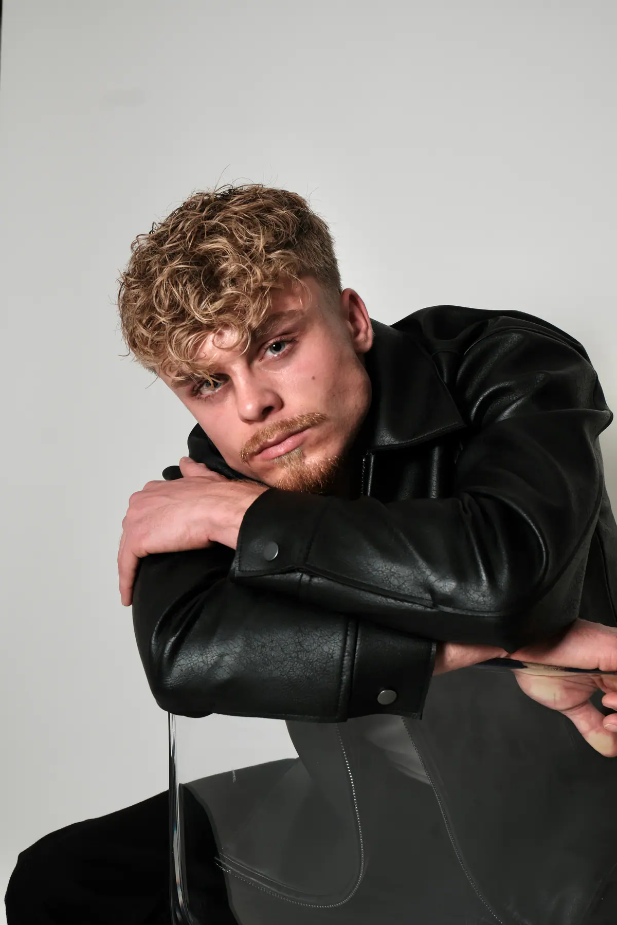 Junger Mann mit blonden Locken in schwarzer Lederjacke, aufgestützt auf einen Acrylstuhl, Studioaufnahme vor hellem Hintergrund