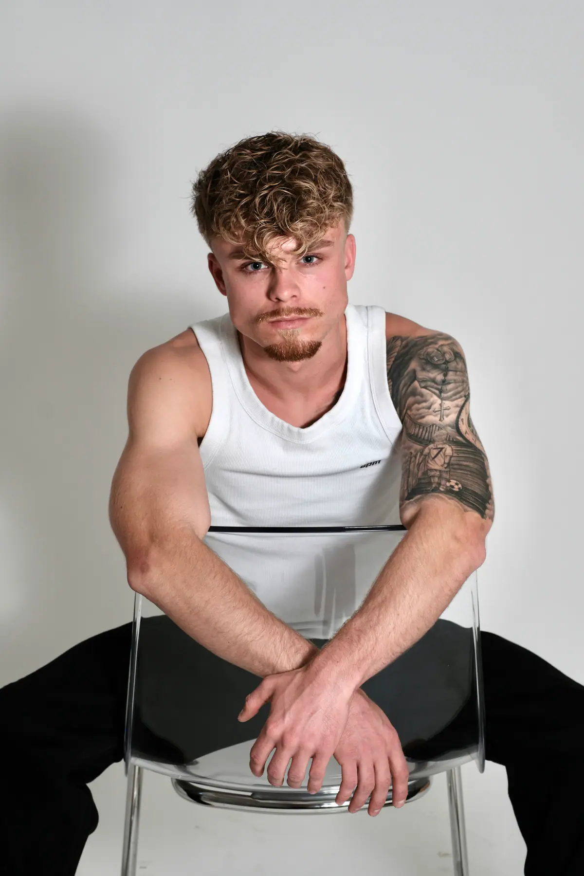 Junger Mann mit blonden Locken und tätowiertem Oberarm sitzt im weißen Tanktop auf einem transparenten Stuhl