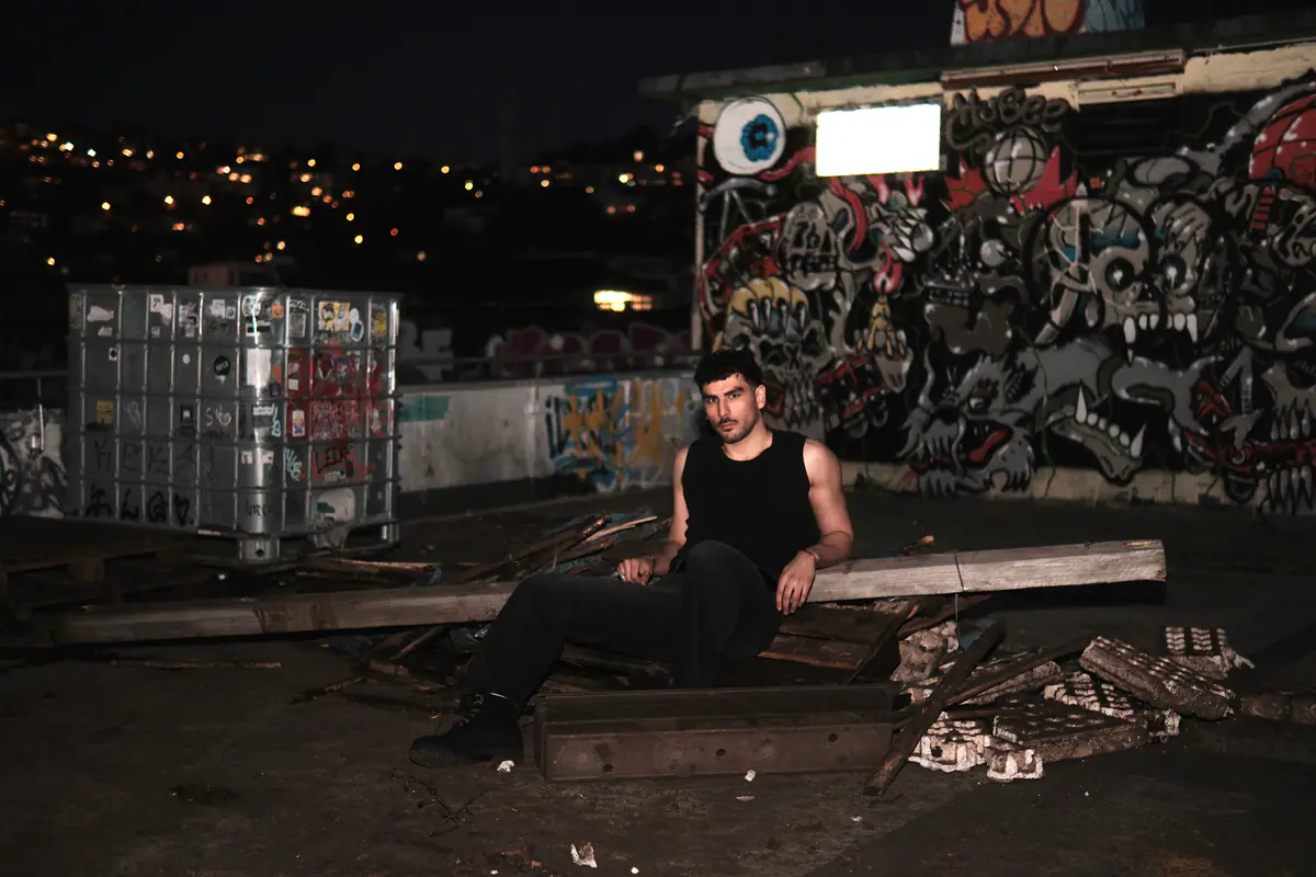 Mann in schwarzem Shirt sitzt nachts auf einem Parkdeck vor einer bunt besprühten Graffitiwand, im Hintergrund die Lichter der Stuttgarter Halbhöhenlage
