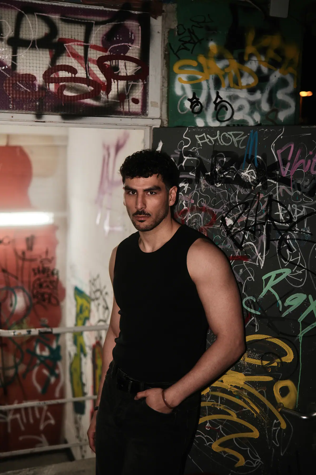 Mann im schwarzen Tanktop steht in einem Treppenhaus vor einer dicht besprühten Graffitiwand