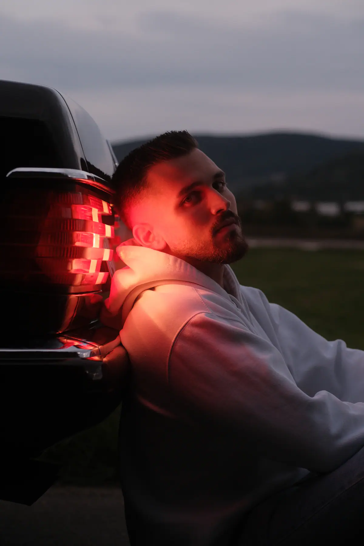 Mann lehnt sich im roten Licht der Rückleuchte über das Heck eines Autos, dahinter die Dämmerung über einem Feld