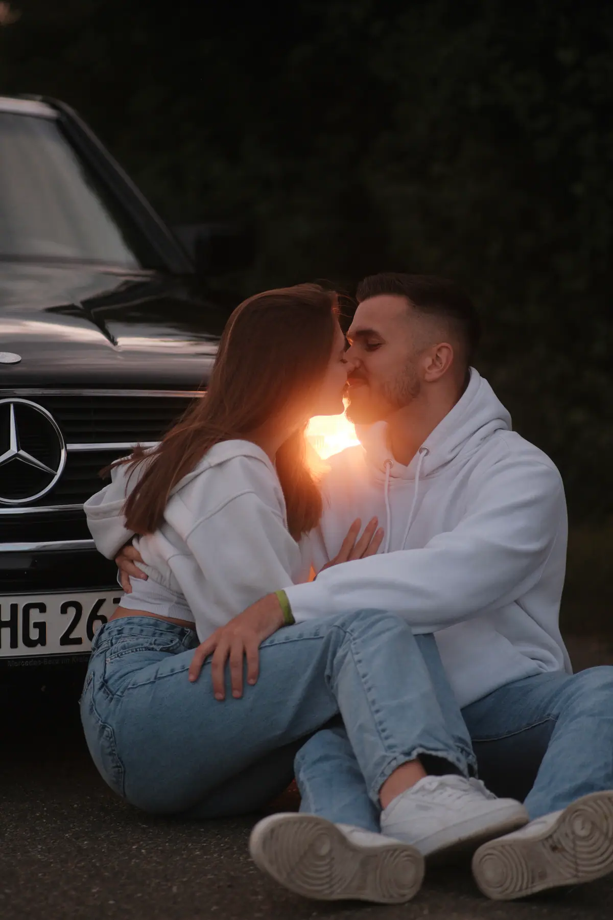 Paar sitzt eng umschlungen vor dem Kühlergrill eines schwarzen Mercedes-Oldtimers, die Sonne steht als Gegenlicht zwischen den beiden